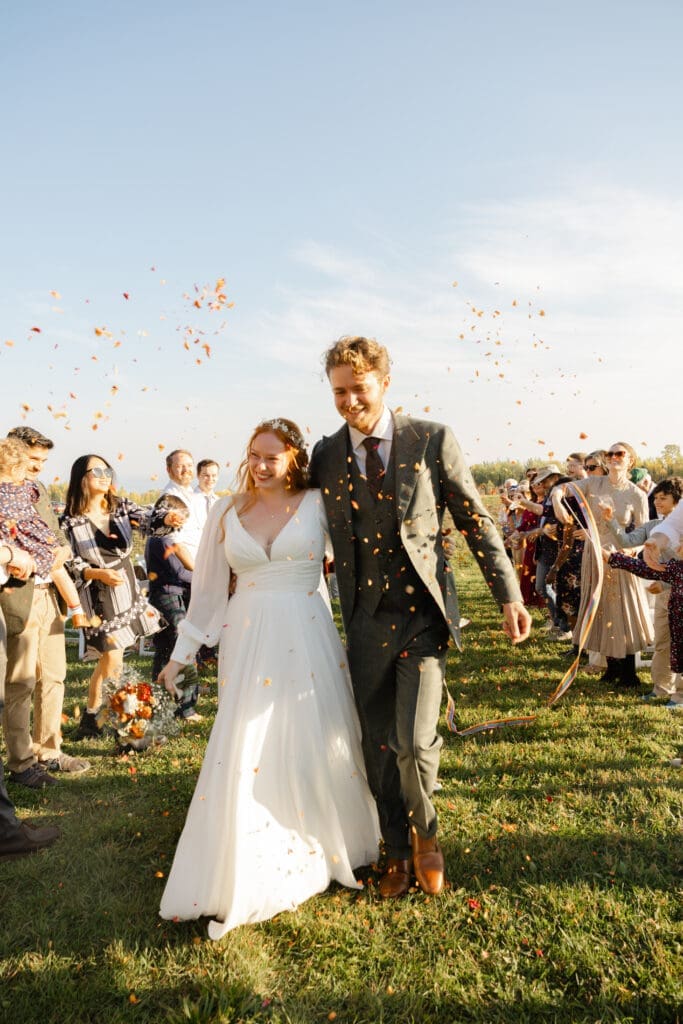 Bride and groom walk hand in hand down an outdoor wedding aisle as guests toss flower petals and confetti during a sunny countryside celebration.